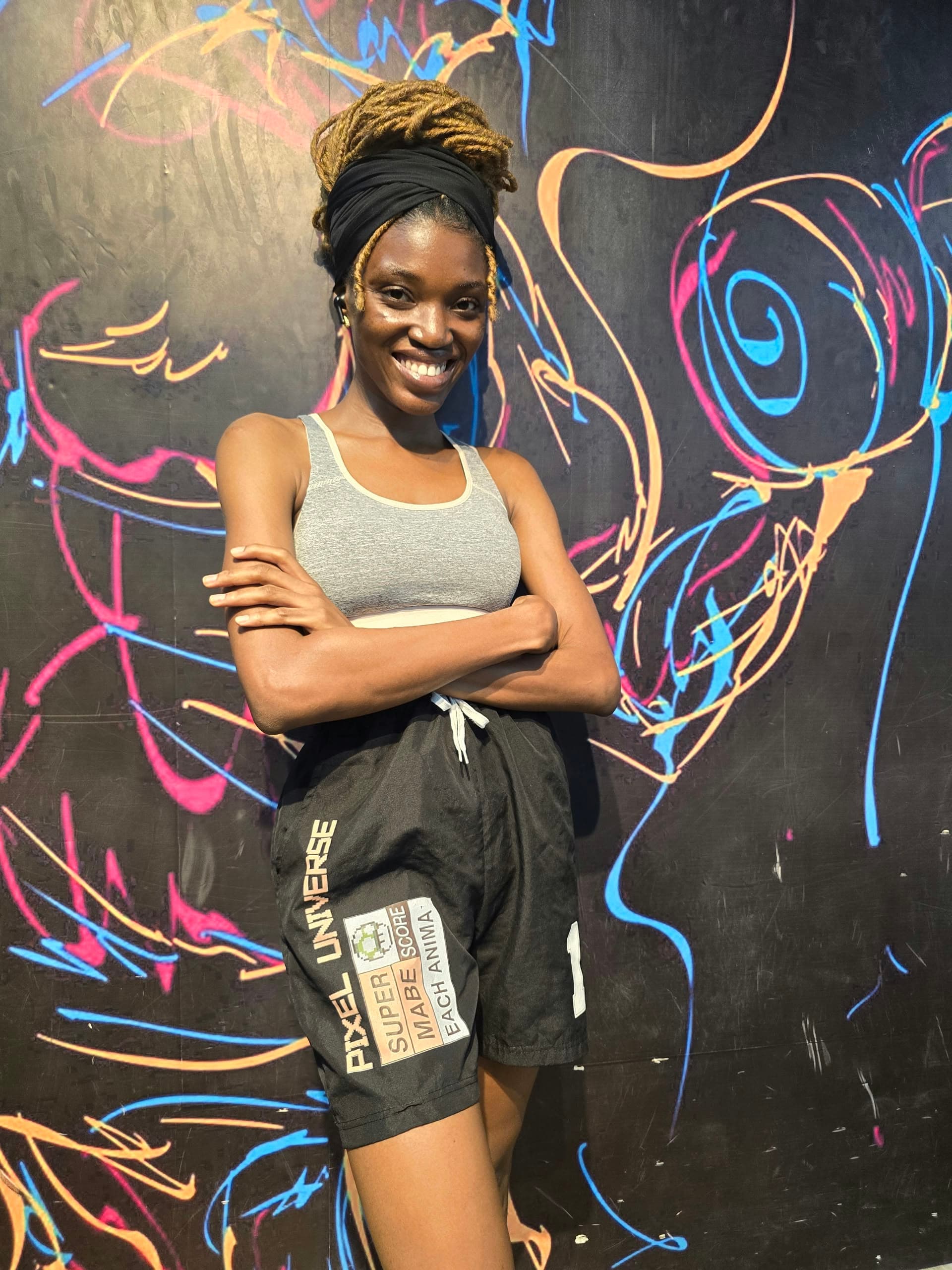 Nimi in athletic wear, smiling with her arms crossed in front of a neon mural